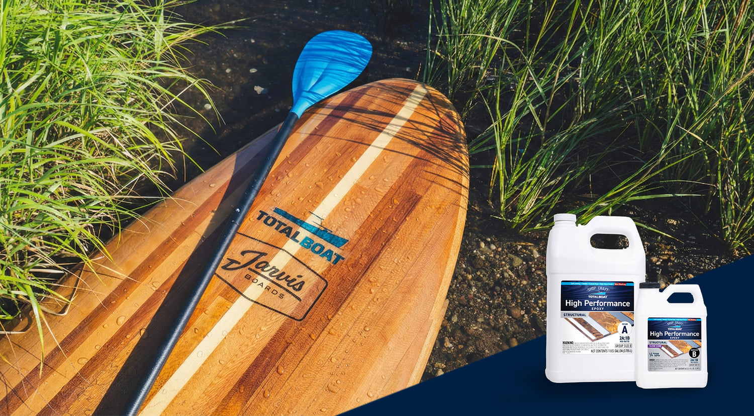 TotalBoat High Performance Epoxy featured on a wooden strip paddle board that's been pulled up onto  the shore. Sea water is beading up on the surface of the paddleboard that's just been out on the water.