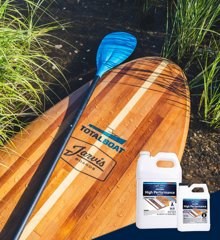 TotalBoat High Performance Epoxy featured on a wooden strip paddle board that's been pulled up onto  the shore. Sea water is beading up on the surface of the paddleboard that's just been out on the water.